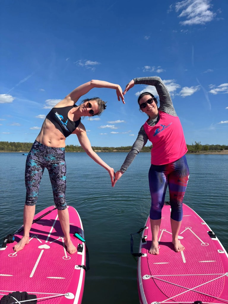 Zwei Teilnehmerinnen formen bei einer SUP-Yoga-Übung gemeinsam ein Herz.