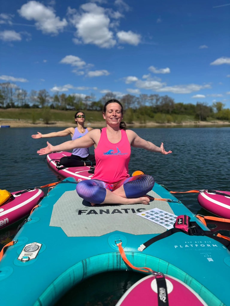 Lisann sitzt mit geöffneten Armen auf einem SUP auf dem See, eine weitere Teilnehmerin übt hinter ihr.